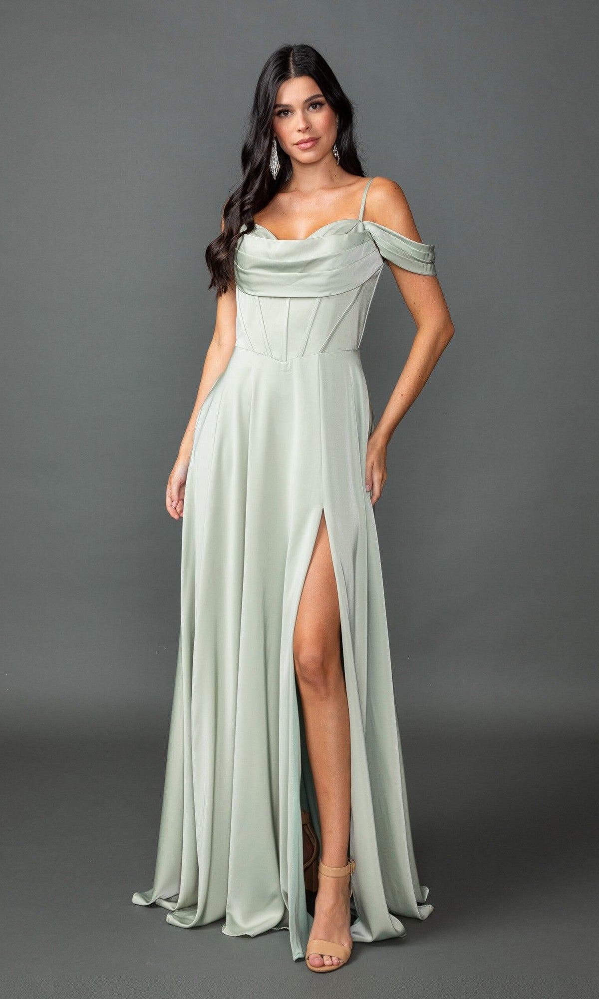 A woman stands against a plain gray background wearing the Long Prom Dress: Dancing Queen 4539, a light sage green off-shoulder A-line gown with a draped corset bodice, thigh-high slit, and high-heeled sandals.